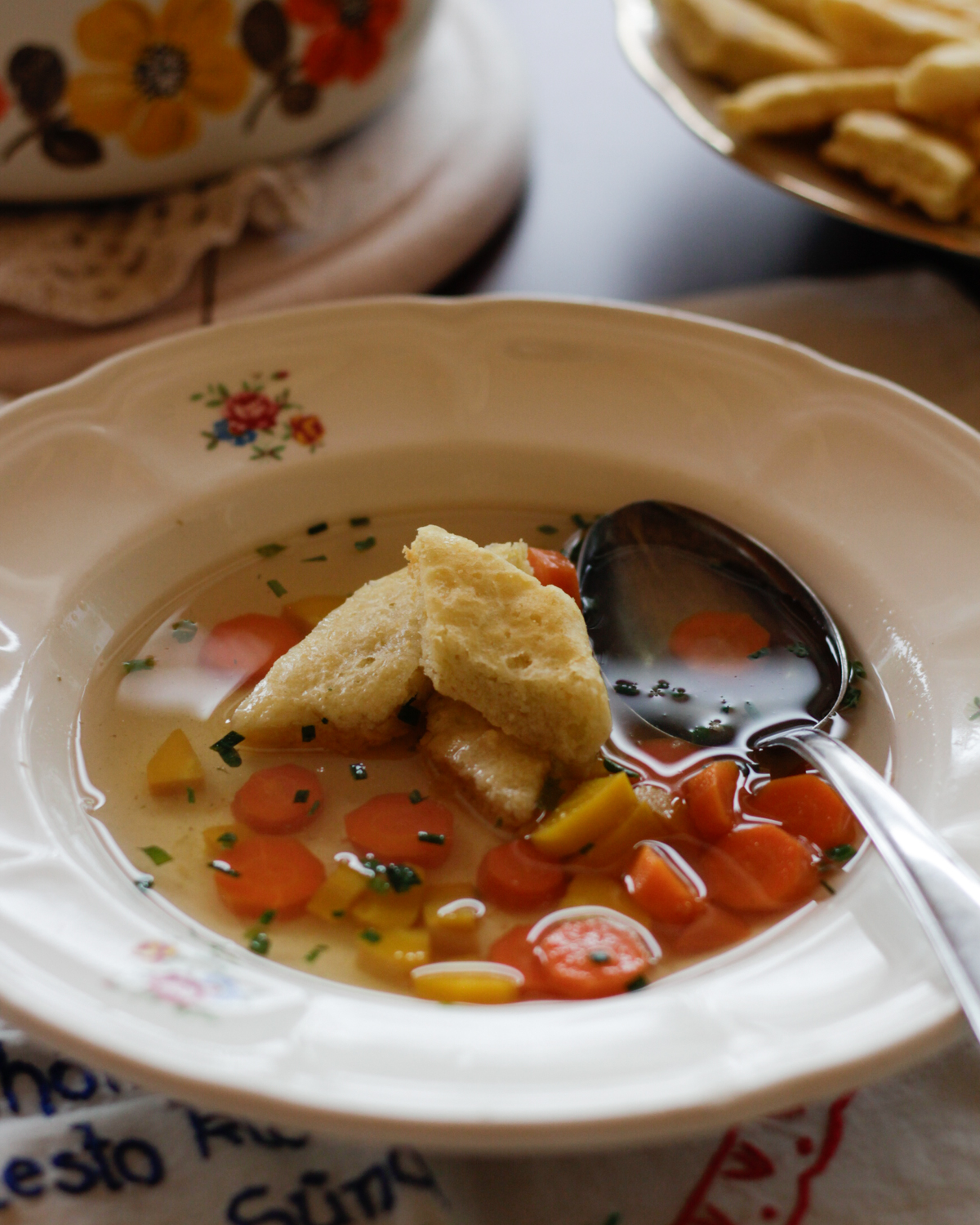 Rautenförmige Biskuitschöberl auf einem blumenverzierten Teller mit  klarer Gemüsesuppe.