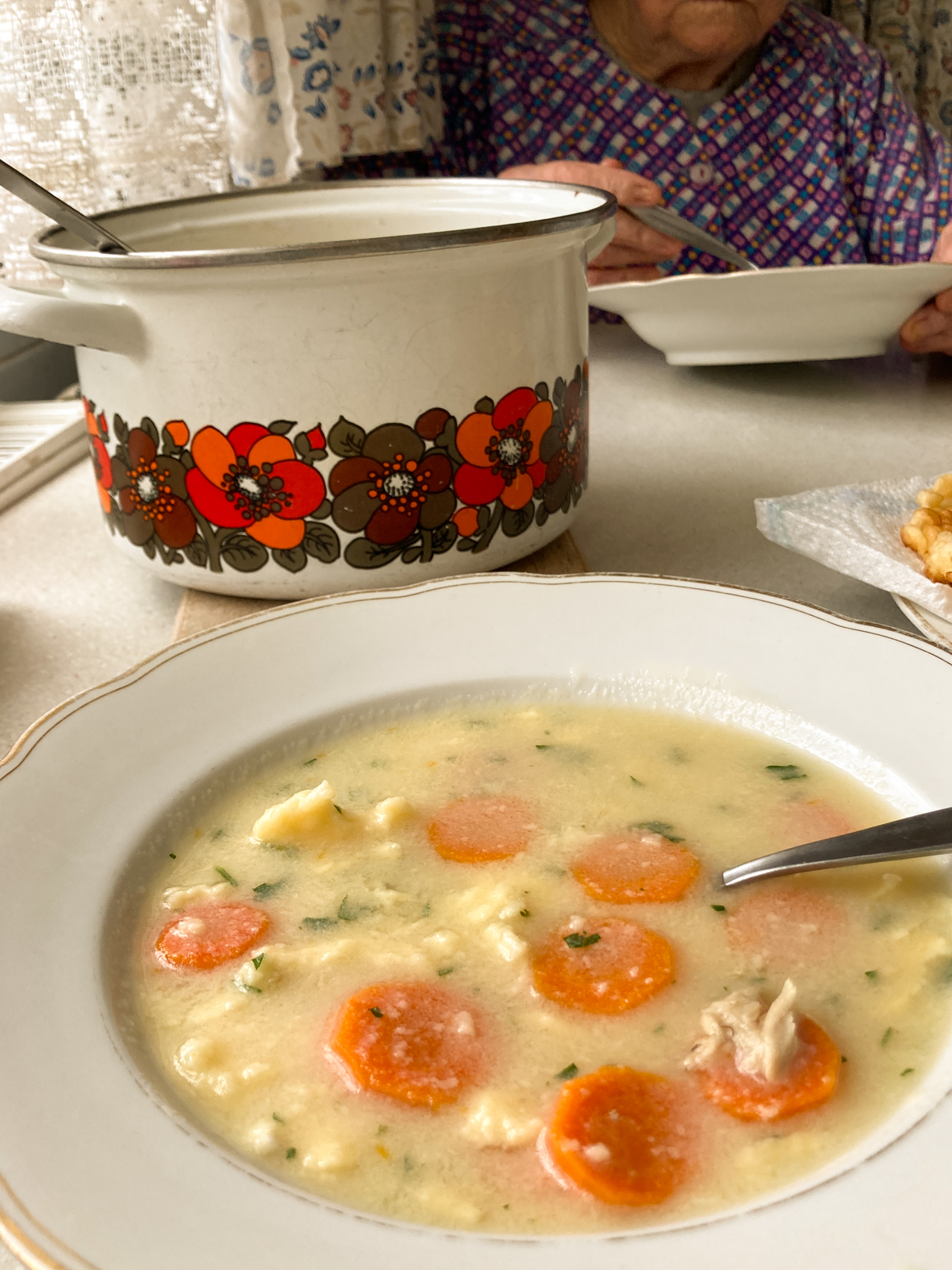 Teller mit Eintropfsuppe und Karottenscheiben, rustikaler Topf und Oma, die Suppe isst im Hintergrund.