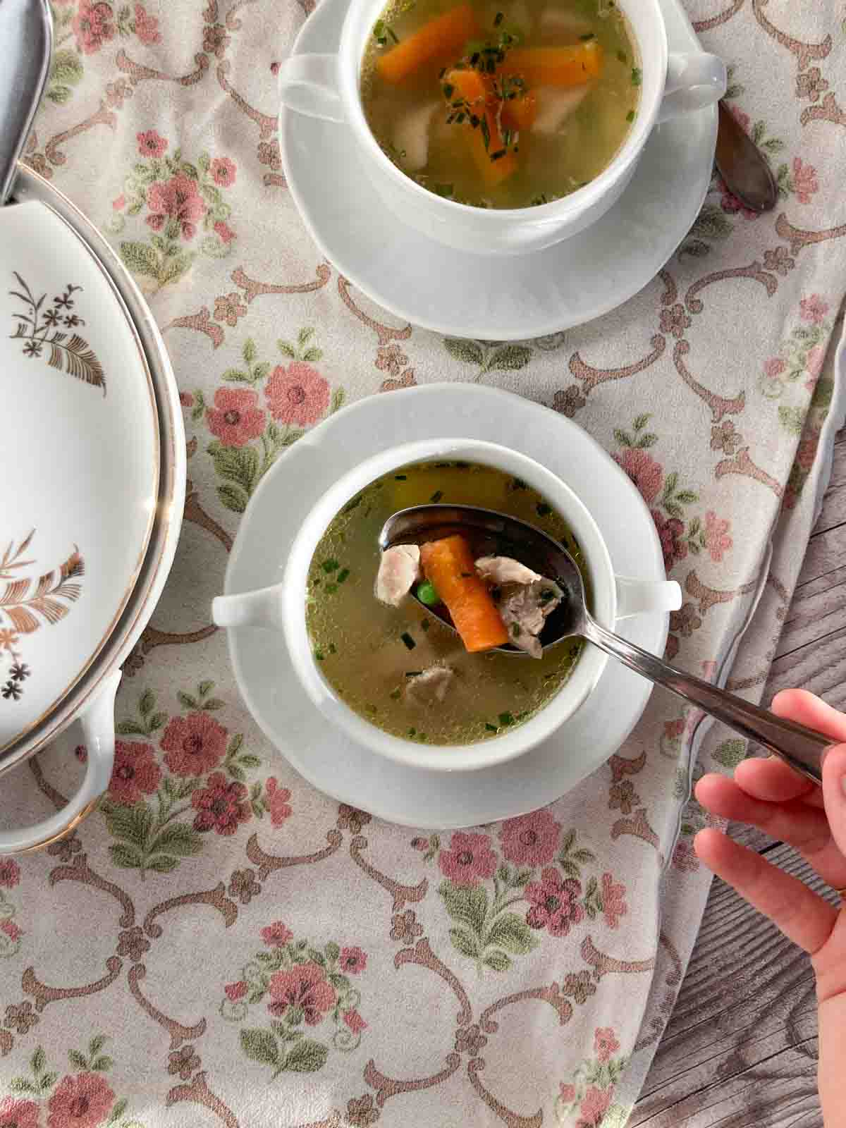 Suppentasse mit Untertasse, gefüllt mit Hühnersuppe, auf rosa geblümtem Tischtuch - easy chicken soup recipe, served in a white soup bowl on a pink flowered table cloth.