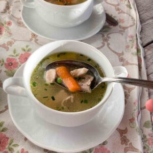 Omas Hühnersuppe serviert in weißer Suppenschüssel mit Henkeln und Untertasse, auf rosa geblümtem Tischtuch - Granny's chicken broth served in small white bowl und pink flower tablecloth.