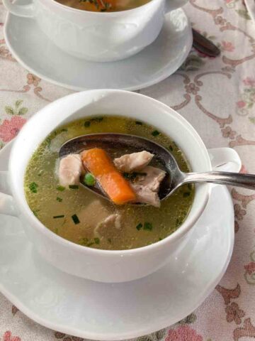 Omas Hühnersuppe serviert in weißer Suppenschüssel mit Henkeln und Untertasse, auf rosa geblümtem Tischtuch - Granny's chicken broth served in small white bowl und pink flower tablecloth.